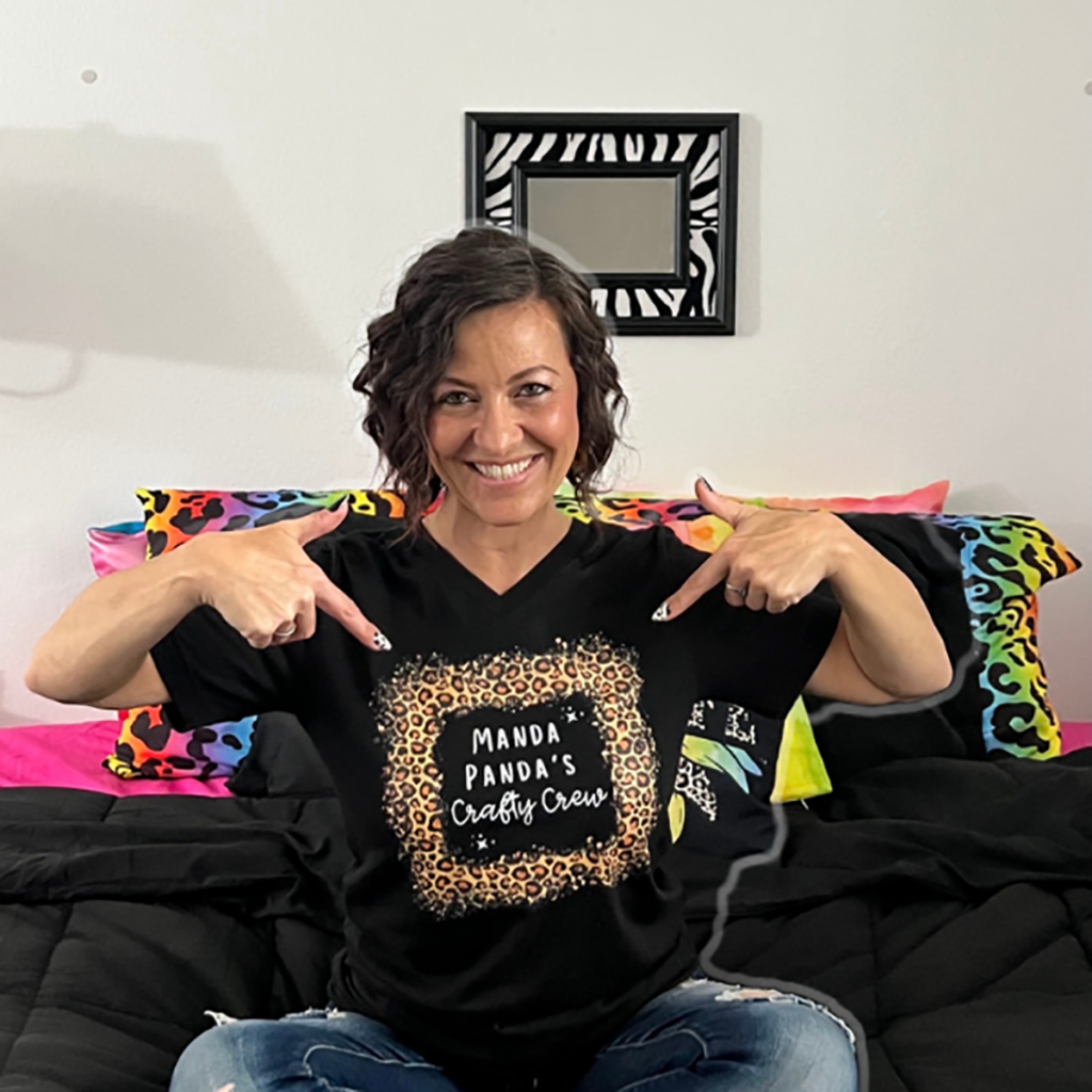 Smiling woman with short dark curly hair sitting on a couch and pointing to the logo on her black "Manda Panda's Crafty Crew" t-shirt.
