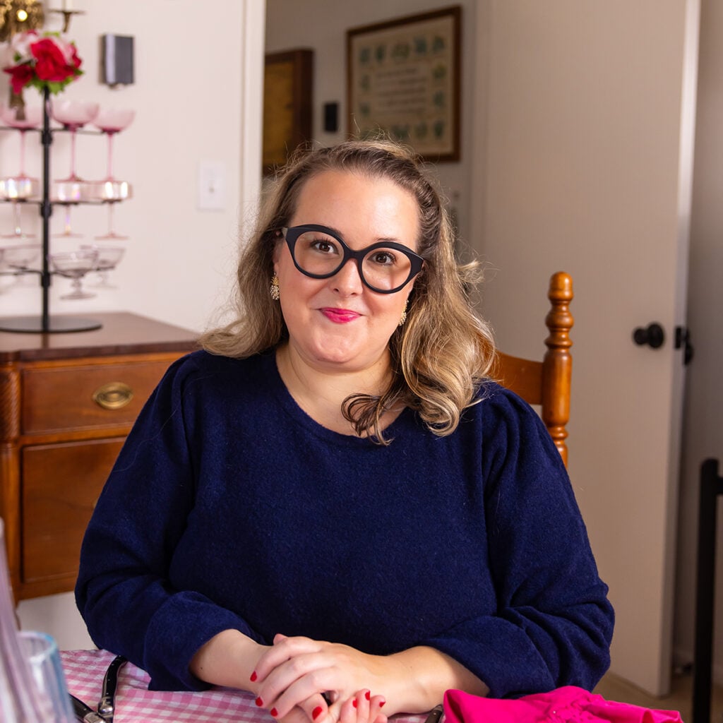 Woman with blonde hair and dark cat-eye glasses smiling while seated at a table in a warmly lit home setting.