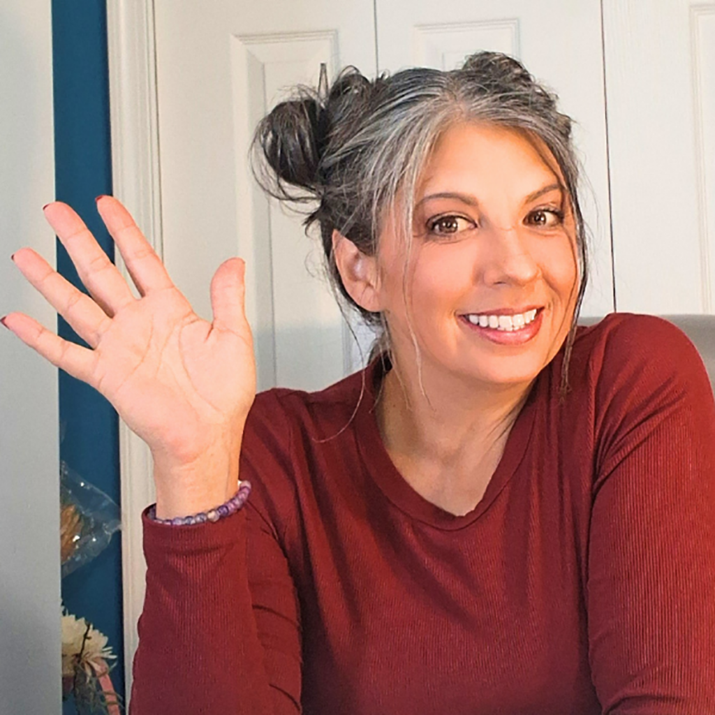 Smiling woman with gray and dark hair styled in two buns, wearing a burgundy top and waving at the camera.