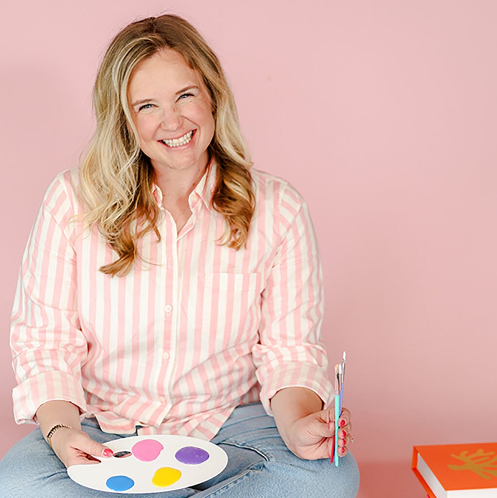 Smiling blonde woman holding a paintbrush and paint palette while seated against a pink background.