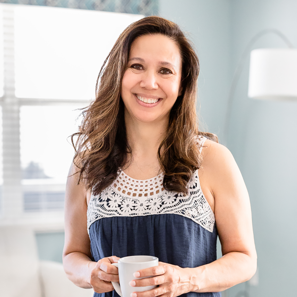 Smiling woman with long dark hair holding a coffee mug in a bright room.