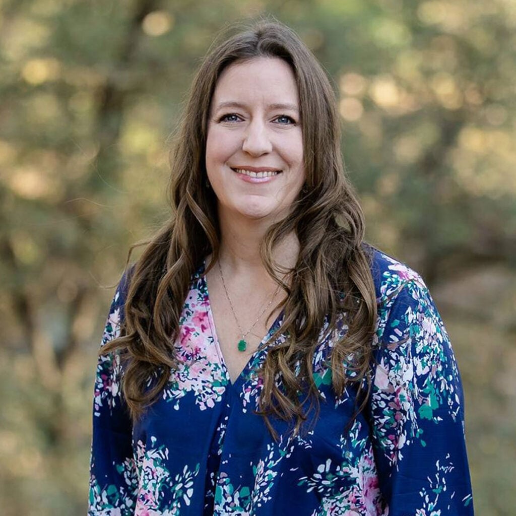 Smiling woman with long brown hair wearing a blue floral blouse outdoors.