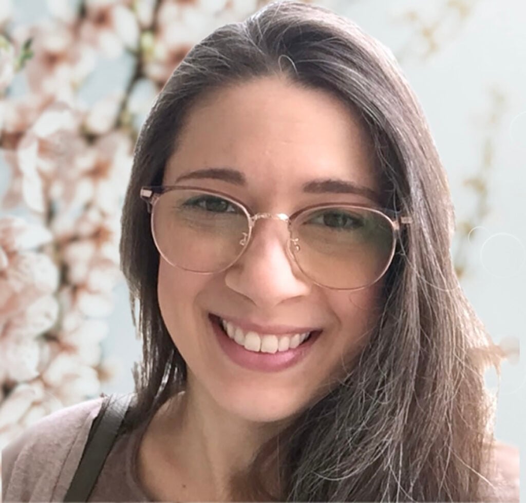 Smiling woman with long dark hair and glasses in front of a soft floral background.