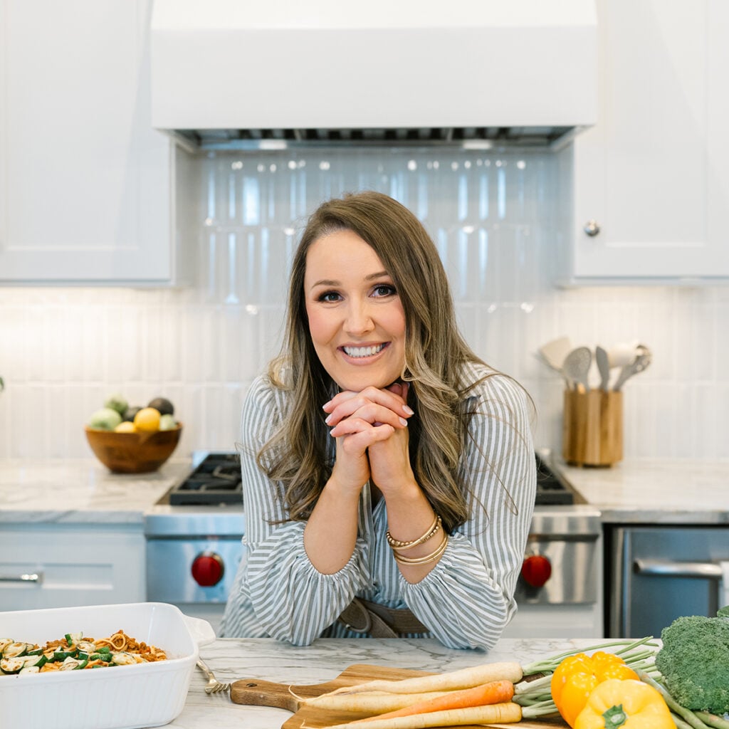 Smiling woman leaning on a kitchen counter with fresh vegetables and a casserole dish nearby.