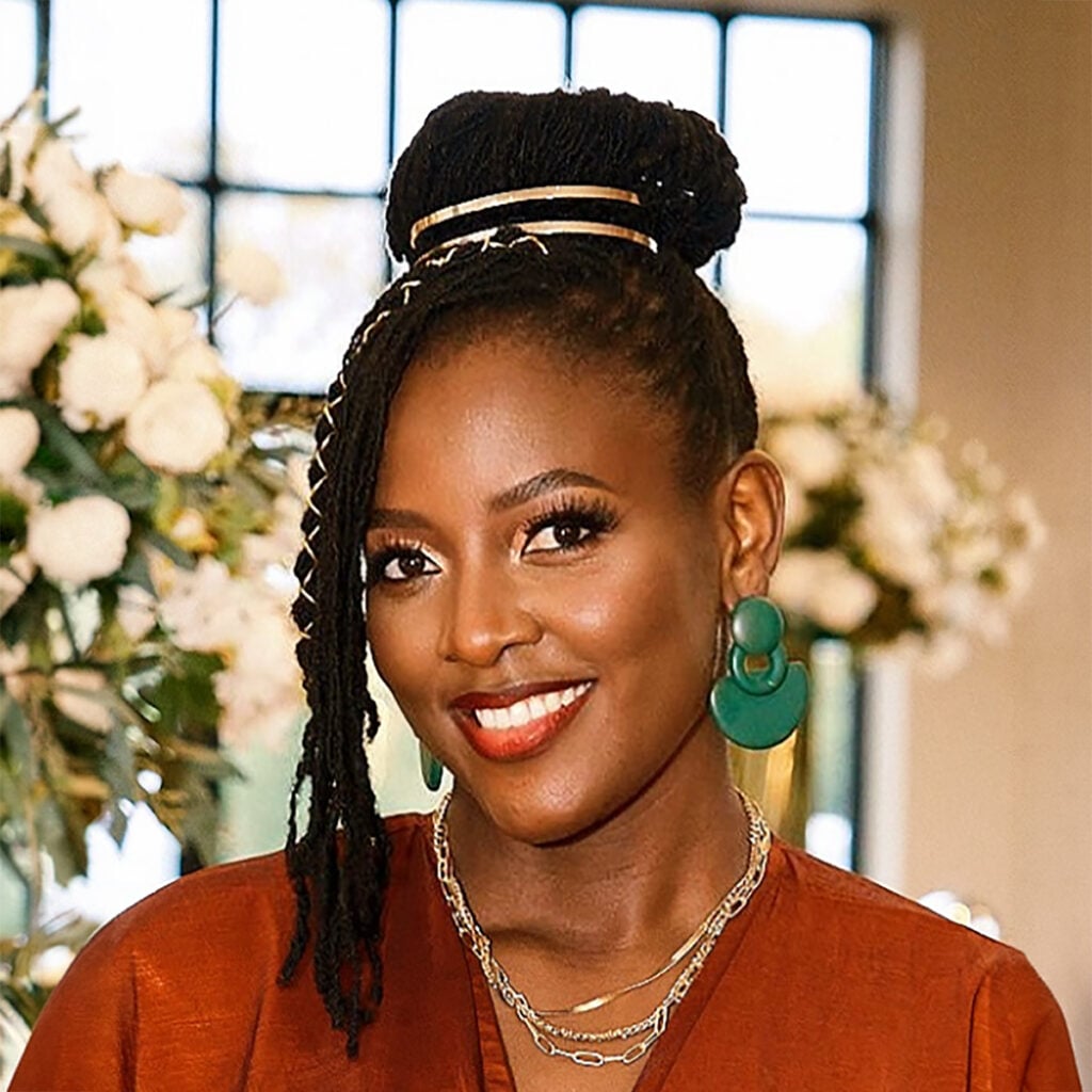 Smiling woman wearing a rust-colored top and green earrings, standing in front of a floral display.