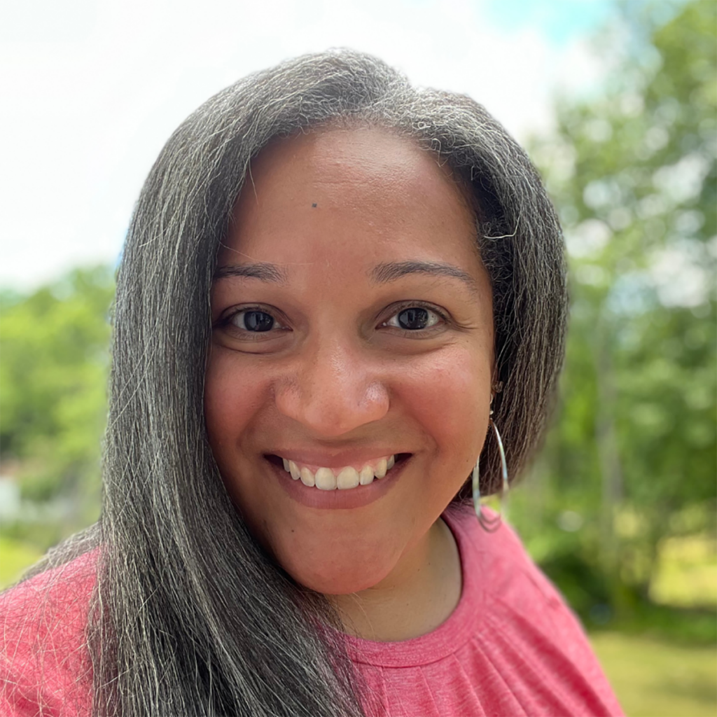 Smiling woman with long dark hair wearing a pink top outdoors with greenery in the background.