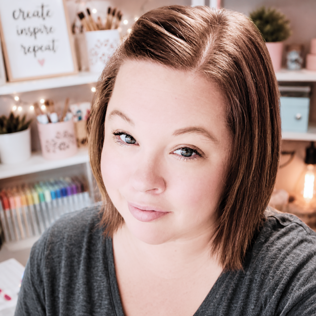 Woman with short brown hair smiling in a creative workspace with art supplies and shelves in the background.