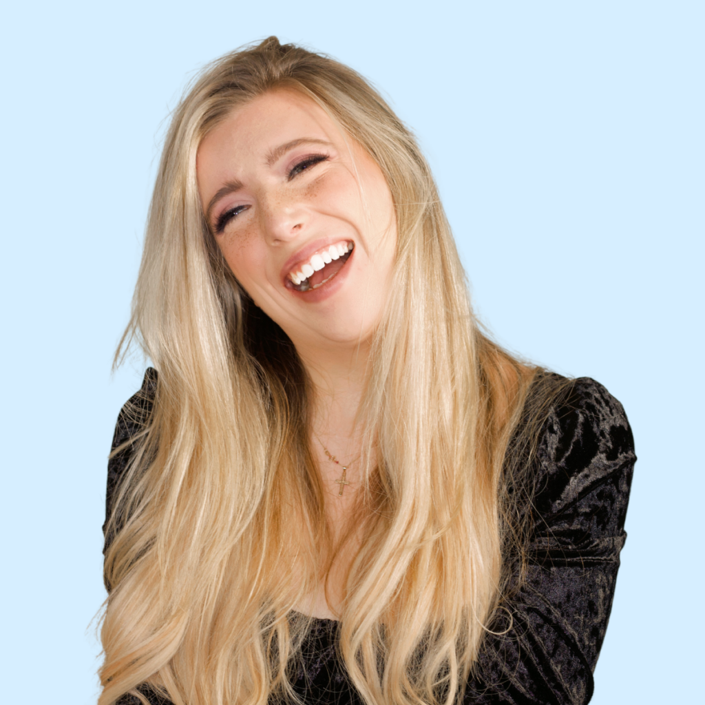 Laughing blonde woman with long hair wearing a black top against a light blue background.