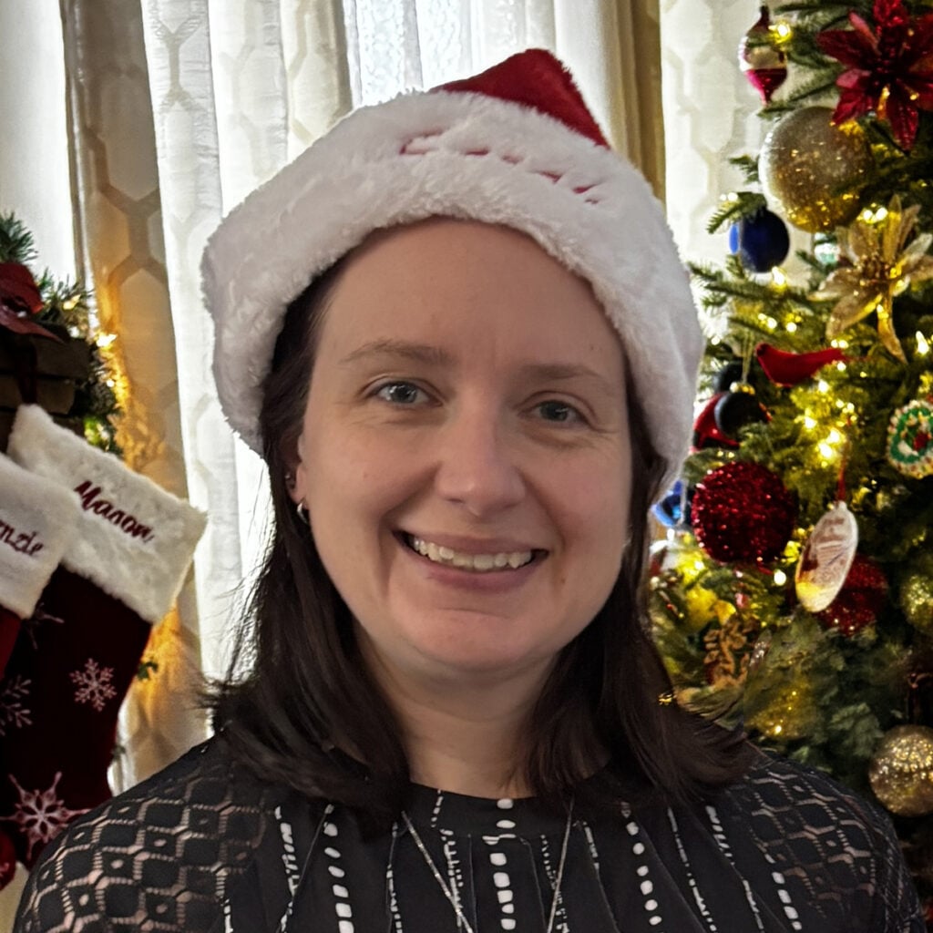 Smiling woman wearing a Santa hat in front of a decorated Christmas tree and holiday stockings.