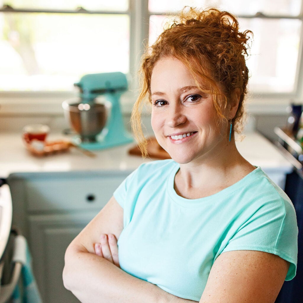 Smiling woman with curly red hair standing in a bright kitchen with arms crossed.